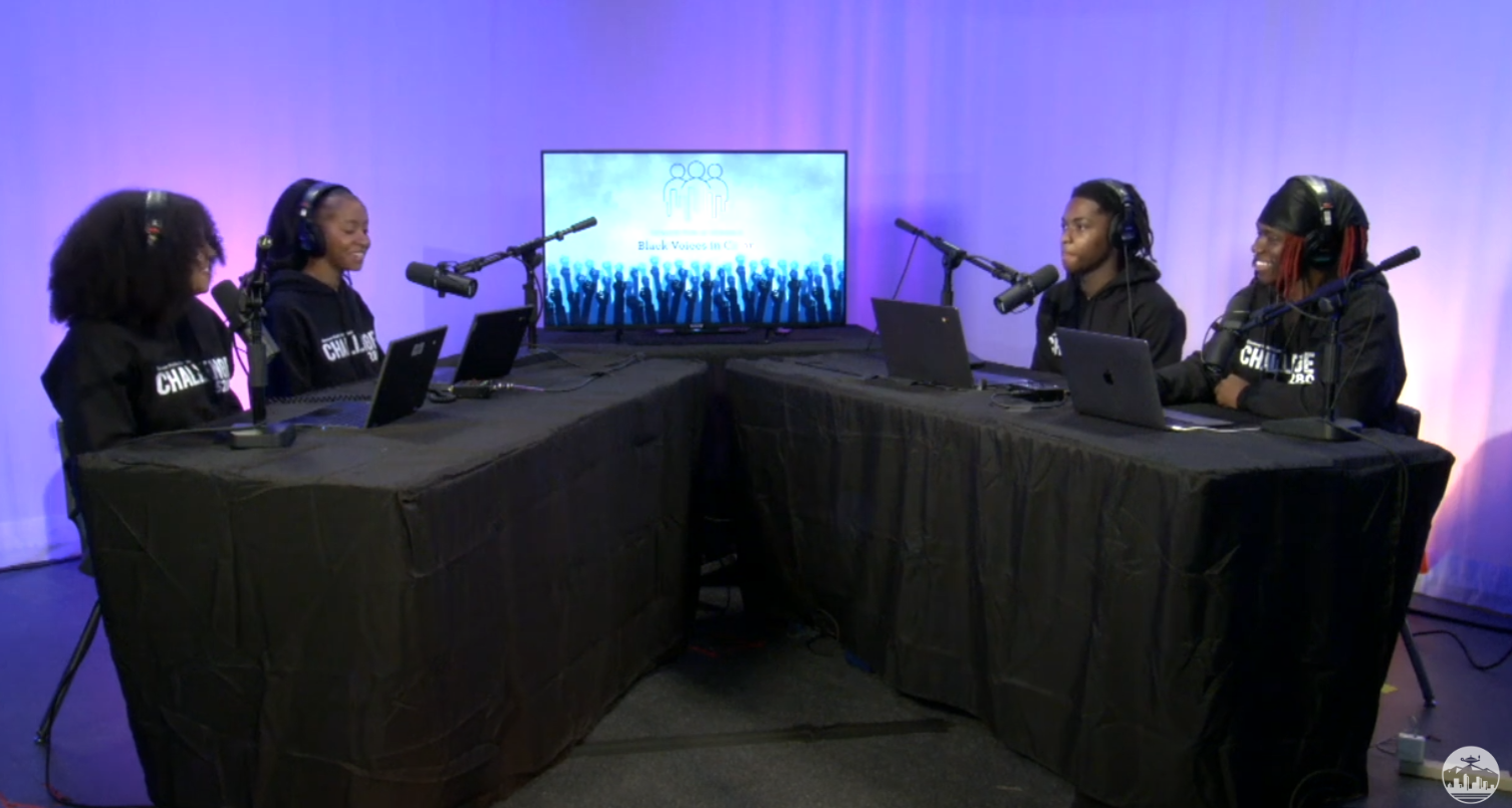 A screenshot from the video feed of the Black Voices in Color Podcast produced by the Denver Public Schools Student Voices Podcast Network. Four students sit at tables facing one another with microphones and laptops.